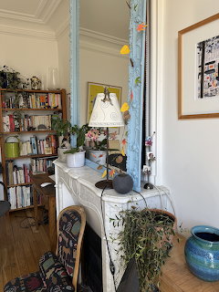 White marble fireplace mantel with plants, lamp, and books in a cozy Parisian apartment with bookshelf and floral mirror