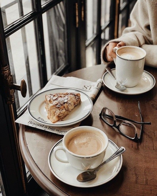Two lattes and an almond croissant on a café table by a window with eyeglasses