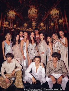 Group of models in white outfits posing in ornate golden ballroom with chandeliers
