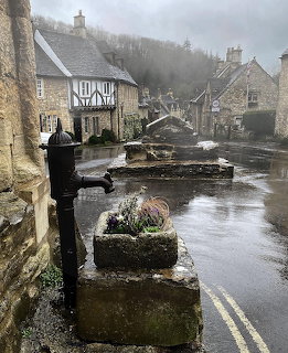 Rainy street view of Castle Combe village with stone cottages and an old water pump in the foreground
