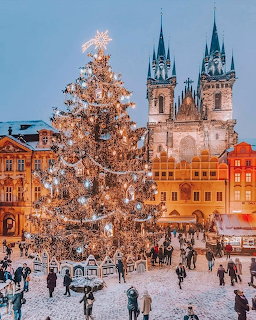 Lit Christmas tree in snowy Prague square at evening with Gothic church towers and people gathered around