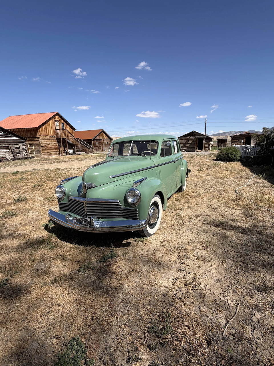 Vintage green 1942 Studebaker Champion parked on dry grass near old wooden buildings