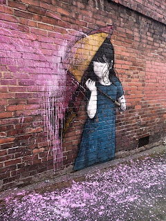 Graffiti mural of a girl with an umbrella on a brick wall with pink paint drips and petals