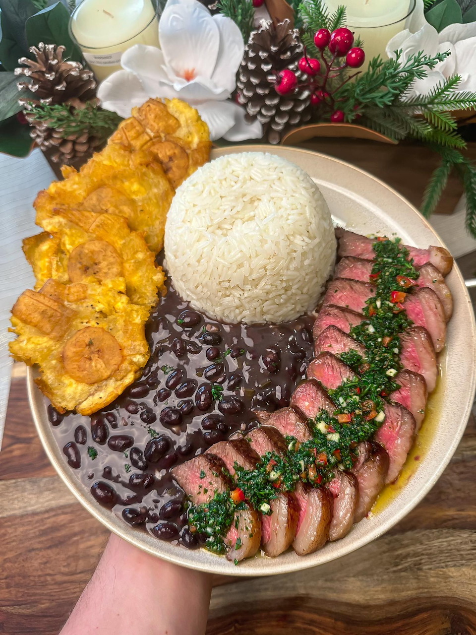 Plate with sliced steak, chimichurri, black beans, white rice, and tostones on a holiday-decorated table