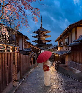 Woman in white kimono with red umbrella walking on wet Kyoto street toward pagoda at twilight