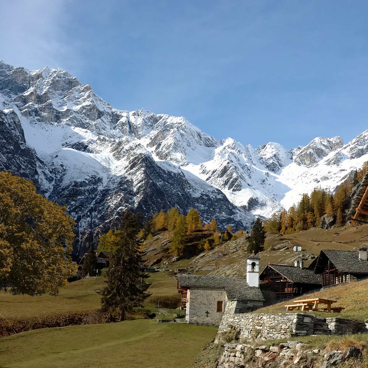 Snow-covered mountain peaks behind a small alpine village with stone houses and green meadows