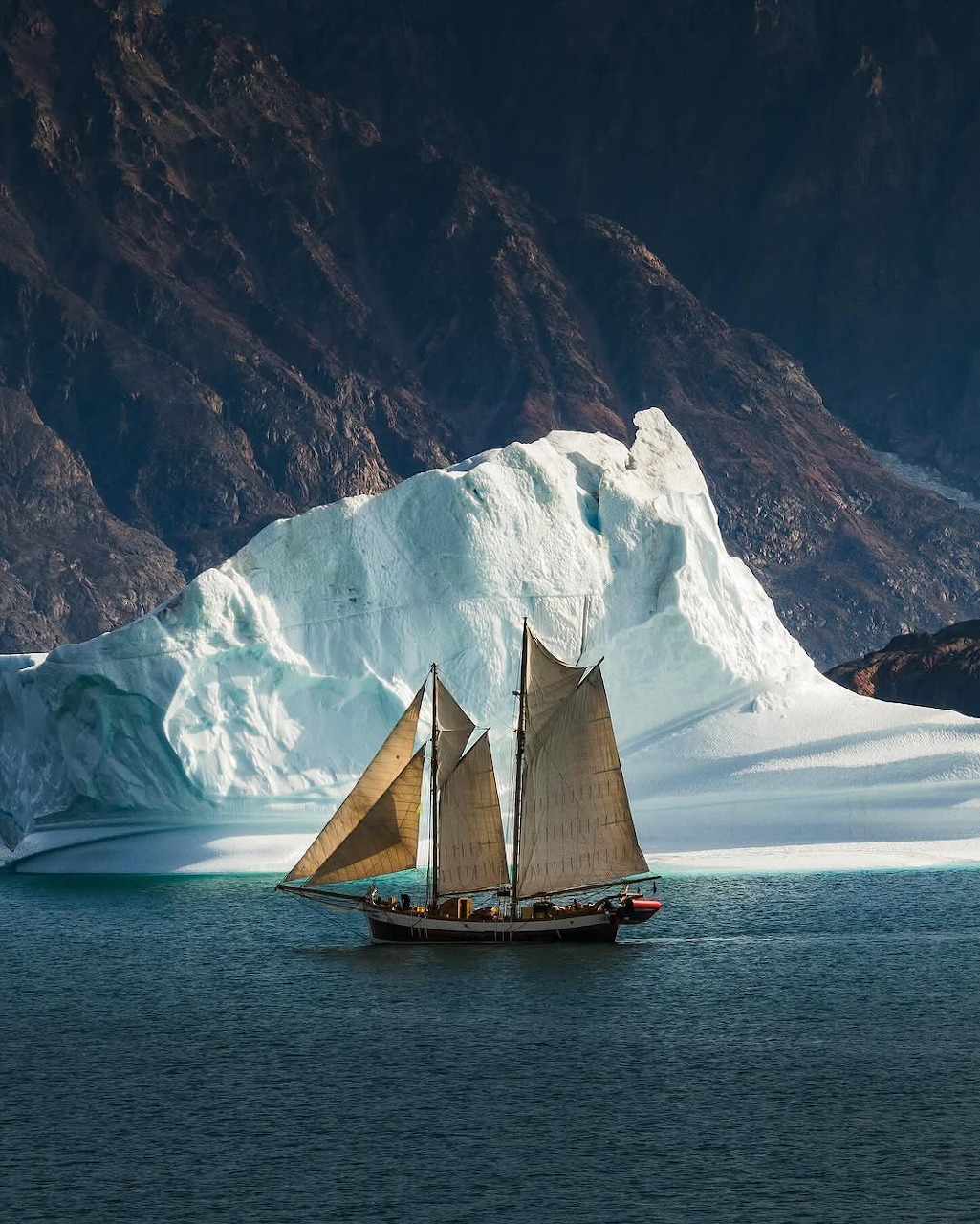 Two-masted sailboat sailing near a large iceberg with rocky mountain cliffs in the background