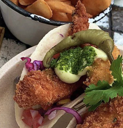 Close-up of crispy fried fish tacos with green sauce, pickled onions, and fries behind