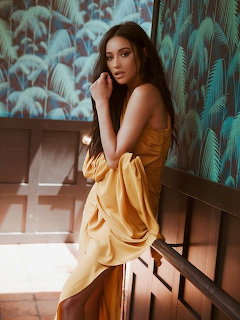 Shay Mitchell in a golden satin gown posing against a tropical leaf wallpaper background