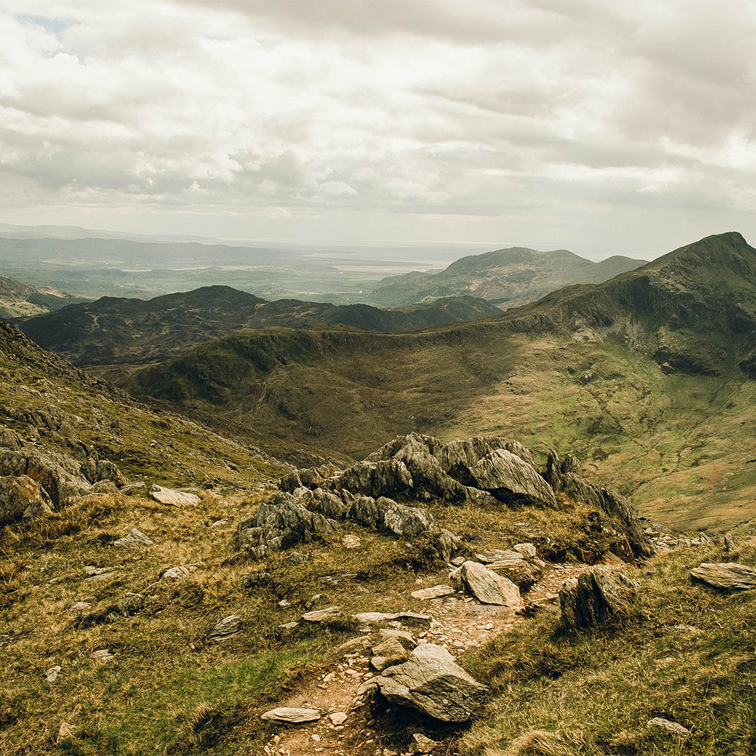 Rocky mountain trail overlooking valleys and peaks under cloudy skies in Snowdonia