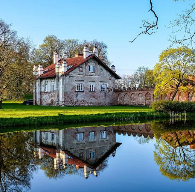 Stone house with red roof reflected in a calm pond beside trees and green grass