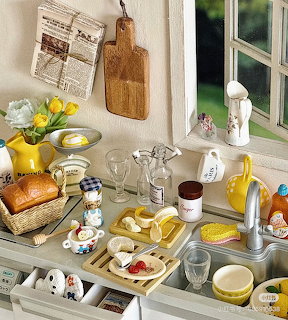 Miniature dollhouse kitchen counter with bread basket, lemons, glassware, yellow tulips, and ceramic items