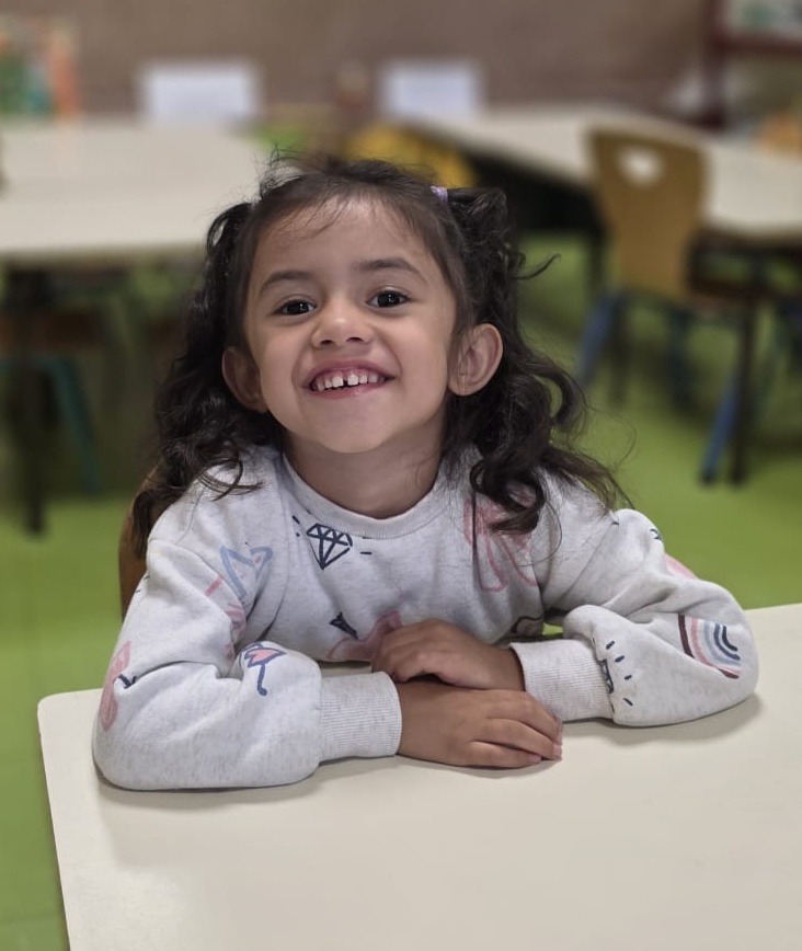Smiling young girl with curly hair sitting at a table in a classroom