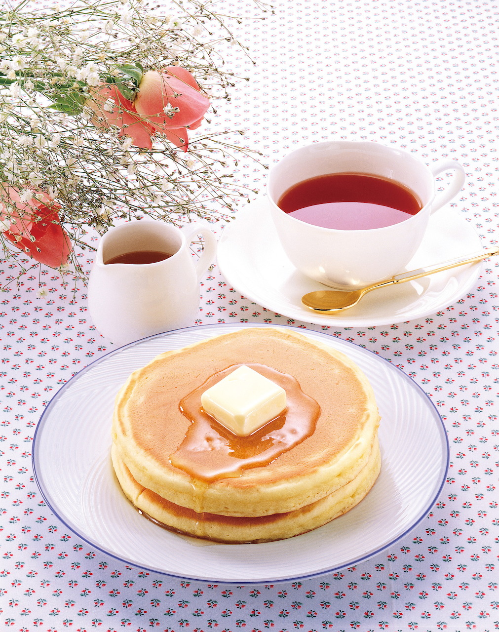 Stack of pancakes with butter and syrup on a plate next to a cup of red tea and a small creamer jug