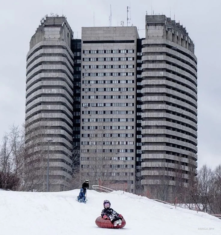 Tall brutalist concrete building with horizontal stripes, winter snow, people sledding in foreground