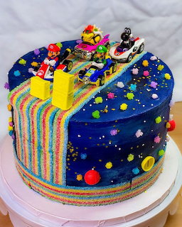 Round layered cake with Super Mario figures, rainbow stripes, yellow pipes, and star decorations on blue frosting