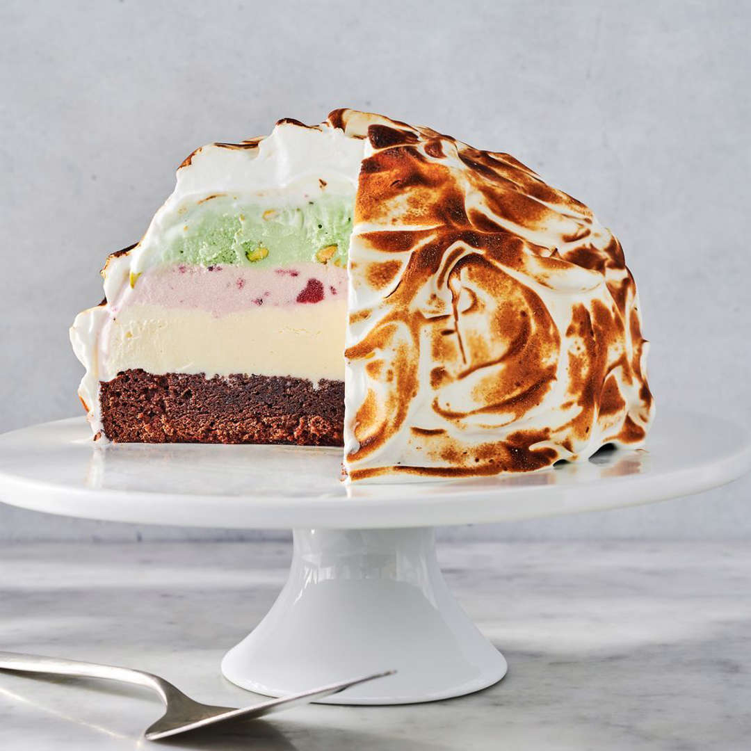 Baked Alaska with golden toasted meringue on white cake stand, showing colorful ice cream layers and chocolate cake base