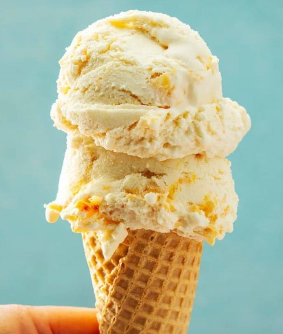 Three scoops of peach ice cream with fruit pieces in a waffle cone held against a teal background