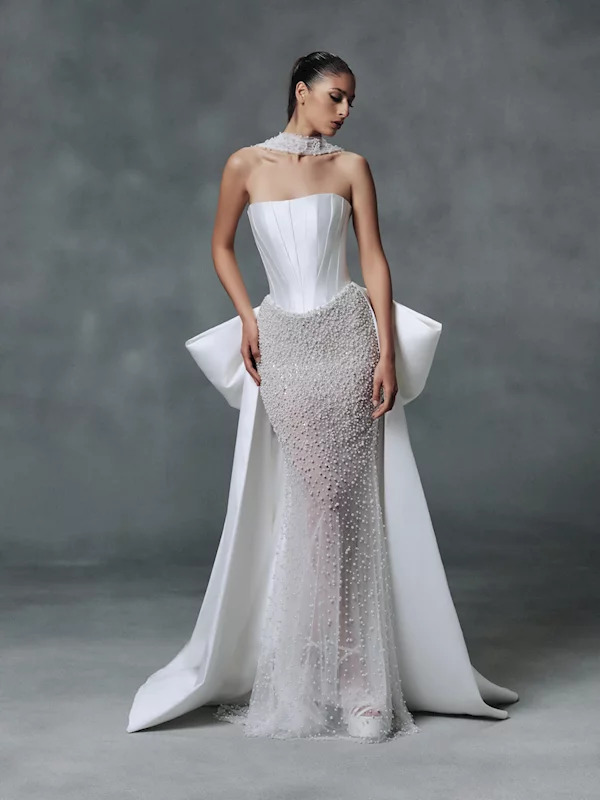 Woman wearing elegant white strapless wedding dress with beaded detailing and flowing fabric against gray background