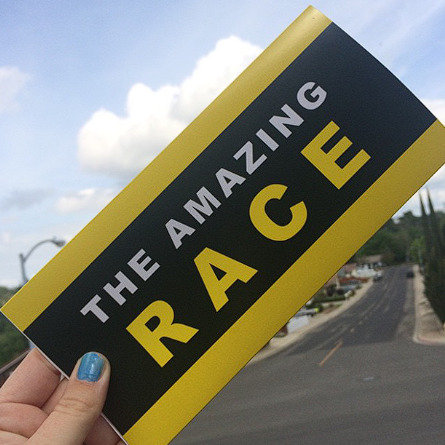 Hand holding a black and yellow The Amazing Race sign above a road