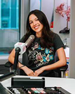 Smiling young woman with long dark hair sitting at a radio studio microphone wearing a black graphic tee