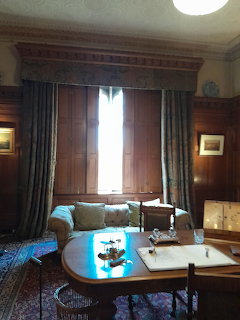 Victorian study with wood paneling, shutered window, sofa, round table, and patterned rug