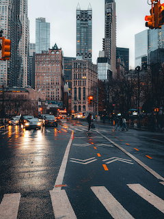 Rainy street intersection in downtown New York City with skyscrapers, traffic lights, and wet pavement reflections.