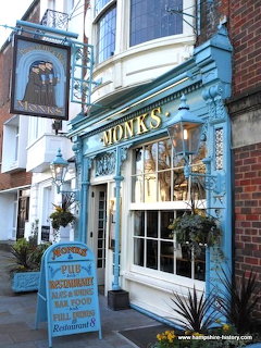 Blue-painted Monks pub with ornate Victorian facade, hanging baskets, and chalkboard sign on an English street
