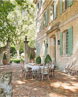 Stone courtyard with wrought iron table and chairs beside a French building with green shutters and trees
