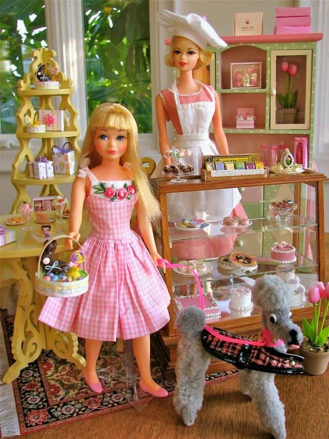 Two blonde dolls in pink dresses standing in a miniature bakery shop with desserts and shelves