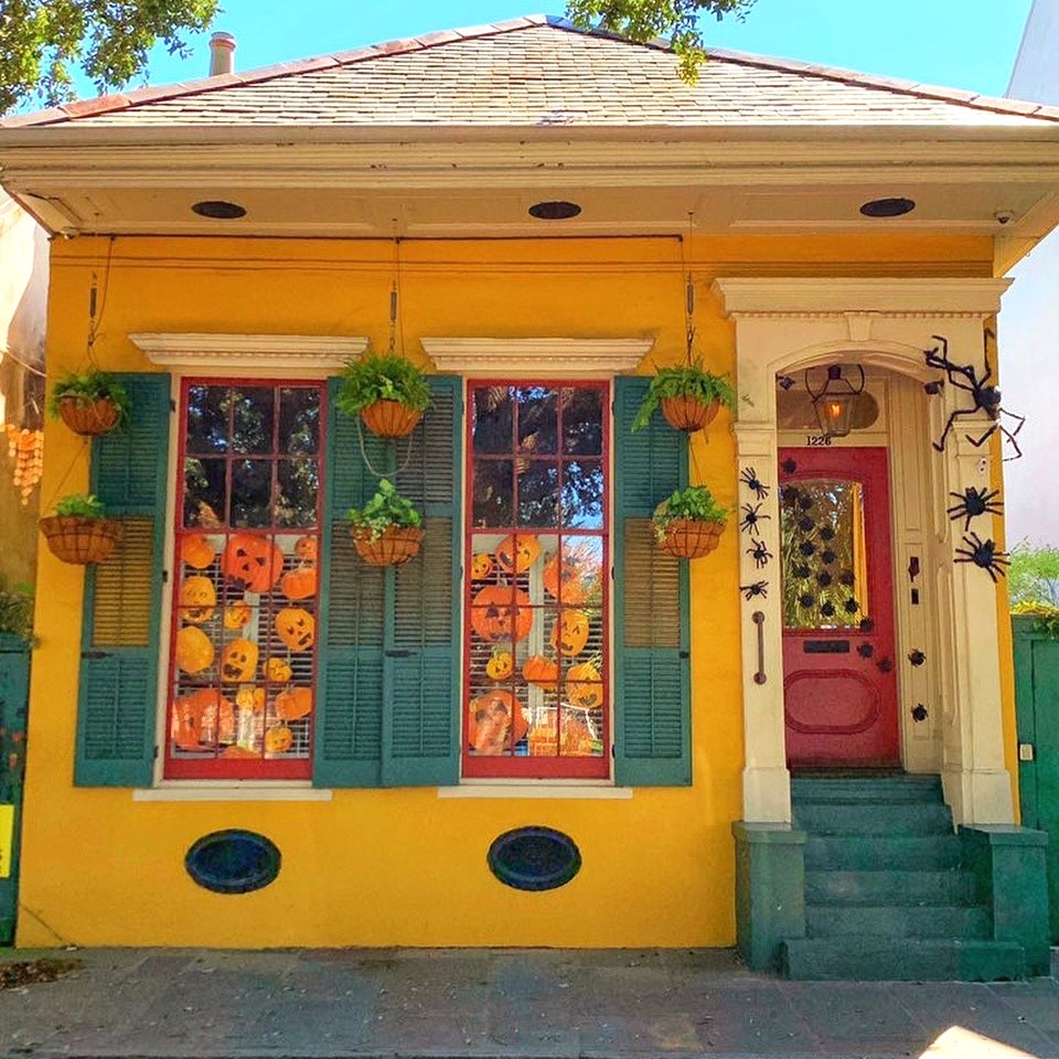 Yellow house with pumpkin window displays, green shuters, hanging baskets, and a red door with Halloween decor