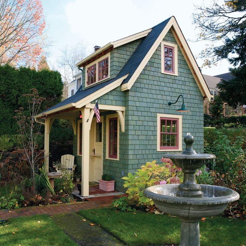 Small blue cottage with peaked roof, red door, and ornate stone fountain in front garden surrounded by greenery