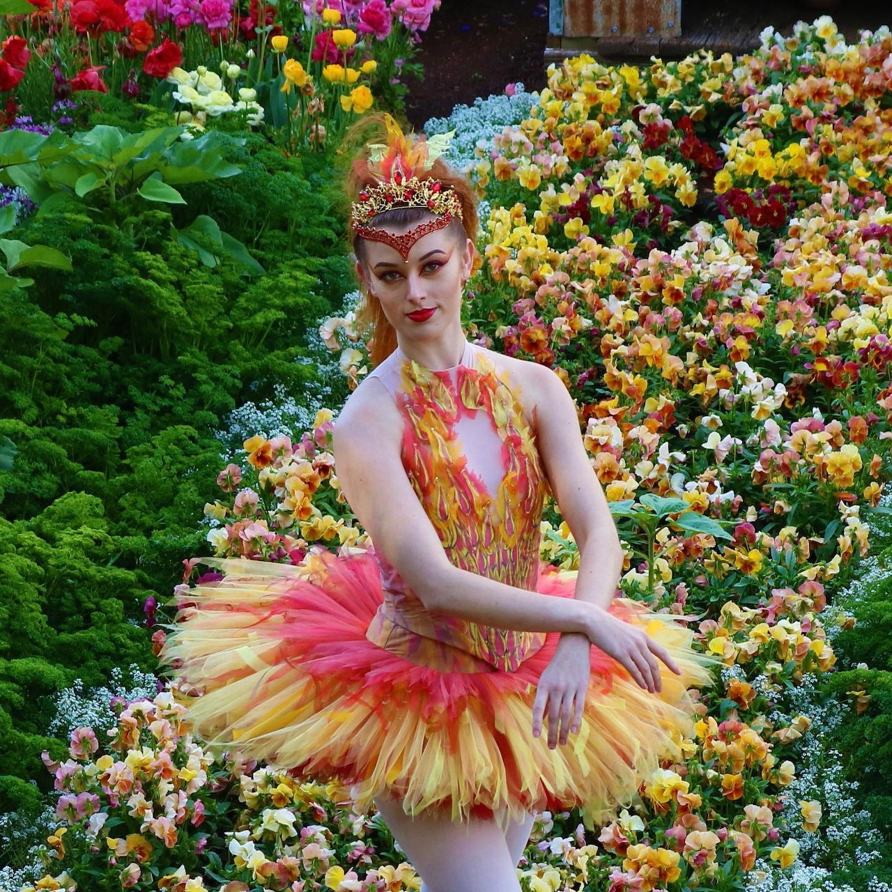Ballet dancer in ornate Firebird costume with golden tutu standing among vibrant garden flowers