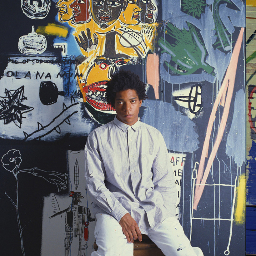 Jean-Michel Basquiat sitting in white clothes in front of a colorful graffiti-style mural