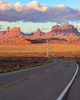 Highway stretching toward Monument Valley red rock buttes at sunset with cloudy sky