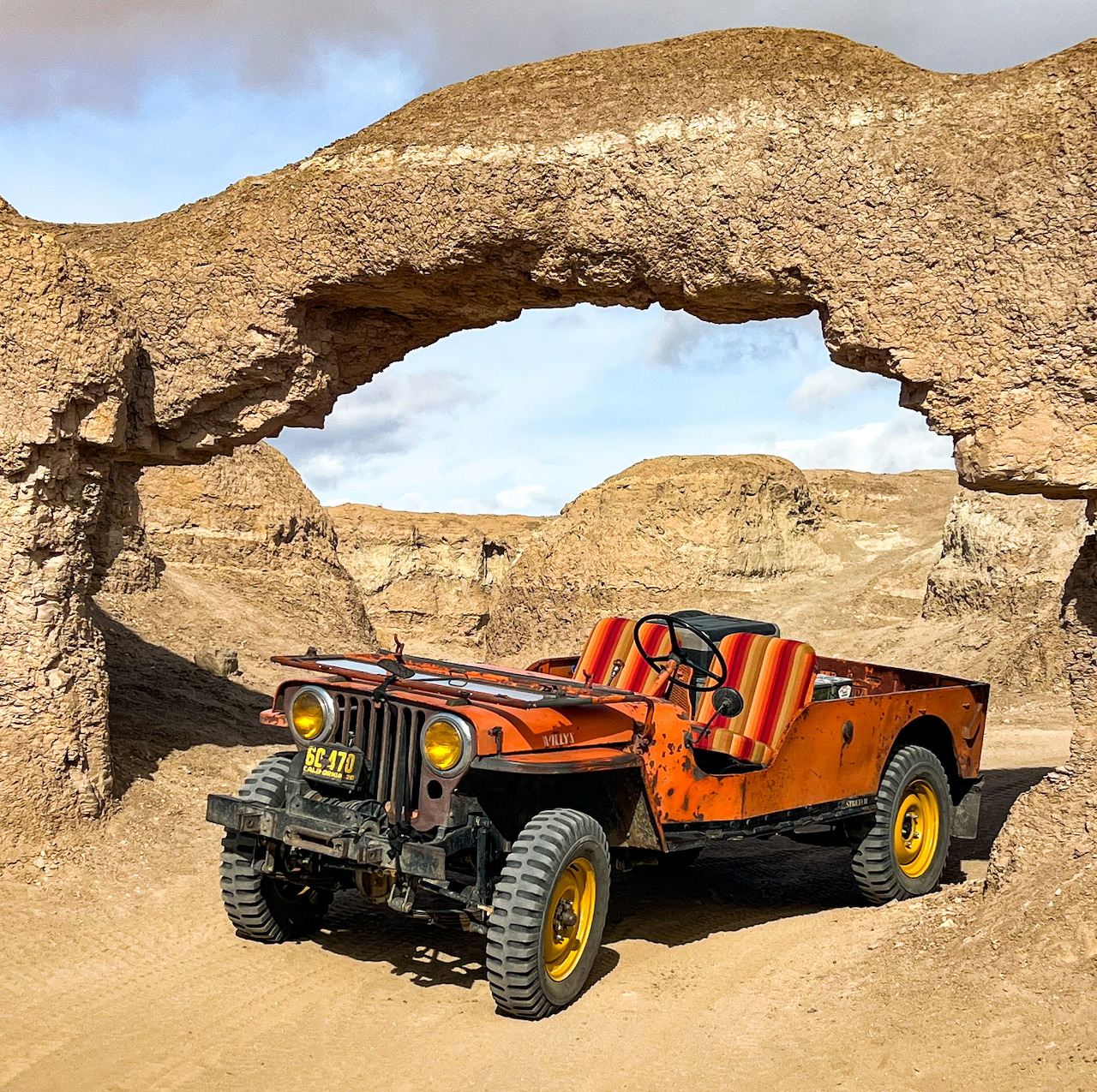 Orange Willys Jeep parked beneath a natural stone arch in a sandy desert setting