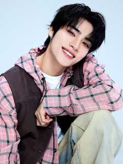 Smiling young man in a pink plaid hoodie and dark vest, seated against a light background.