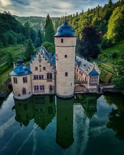 White castle with blue roofs and round tower reflected in surrounding moat water, aerial view with forest