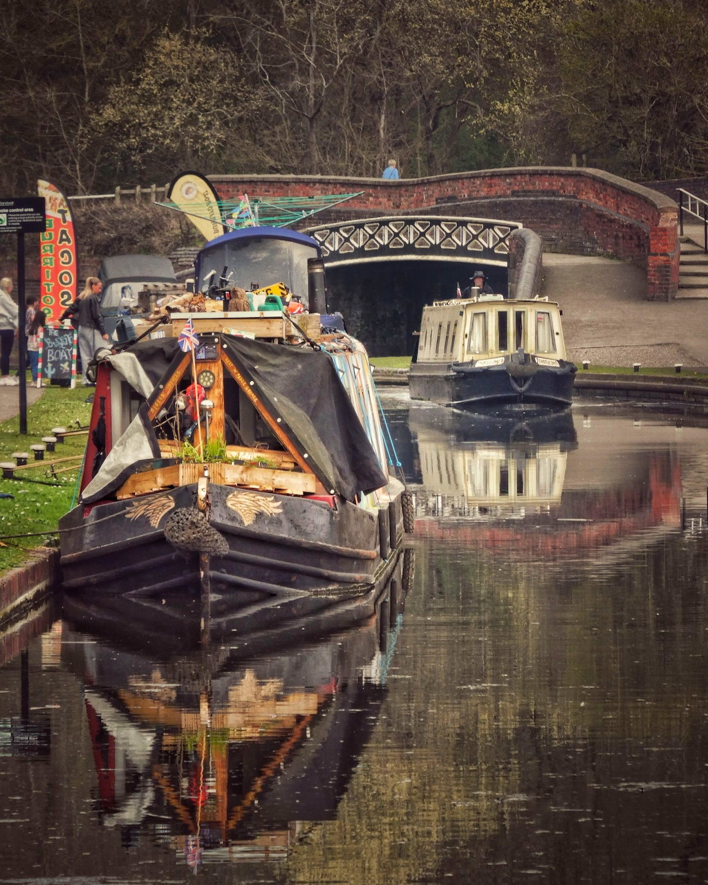 Narrowboats moored on a calm canal with reflections and a red brick arched bridge in background