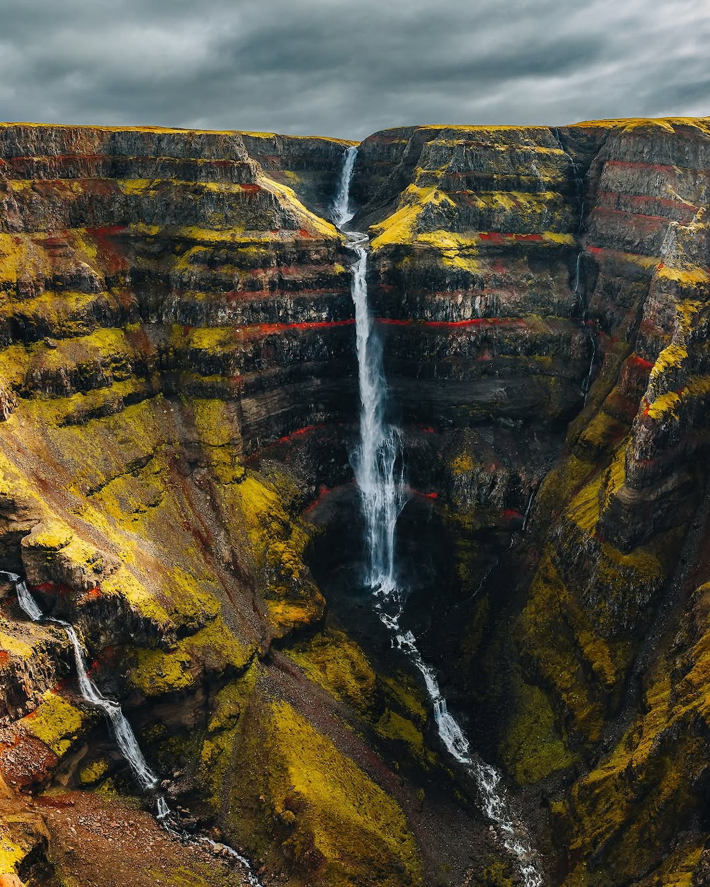 Aerial view of a tall waterfall cascading through a deep rocky canyon with dark stormy clouds above.