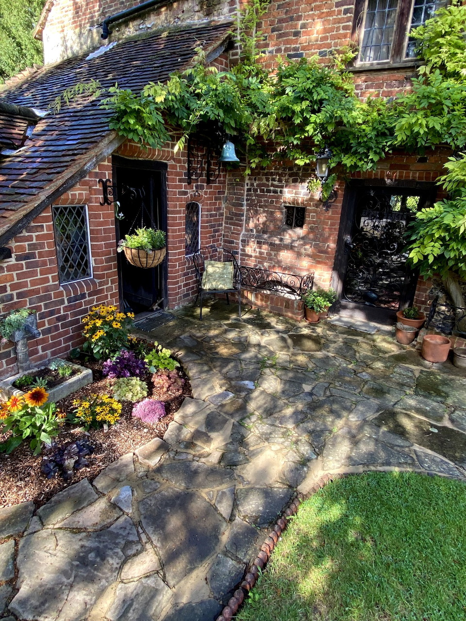 Brick cottage with climbing vines, stone patio, and colorful potted flowers in a garden setting