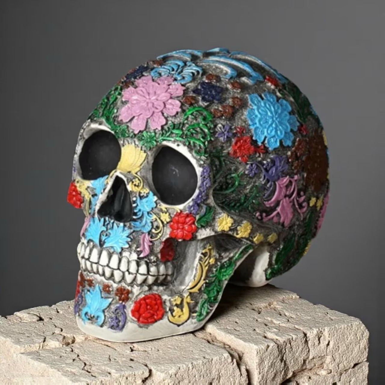 Colorful floral-patterned skull sculpture on a rough stone pedestal against a gray background