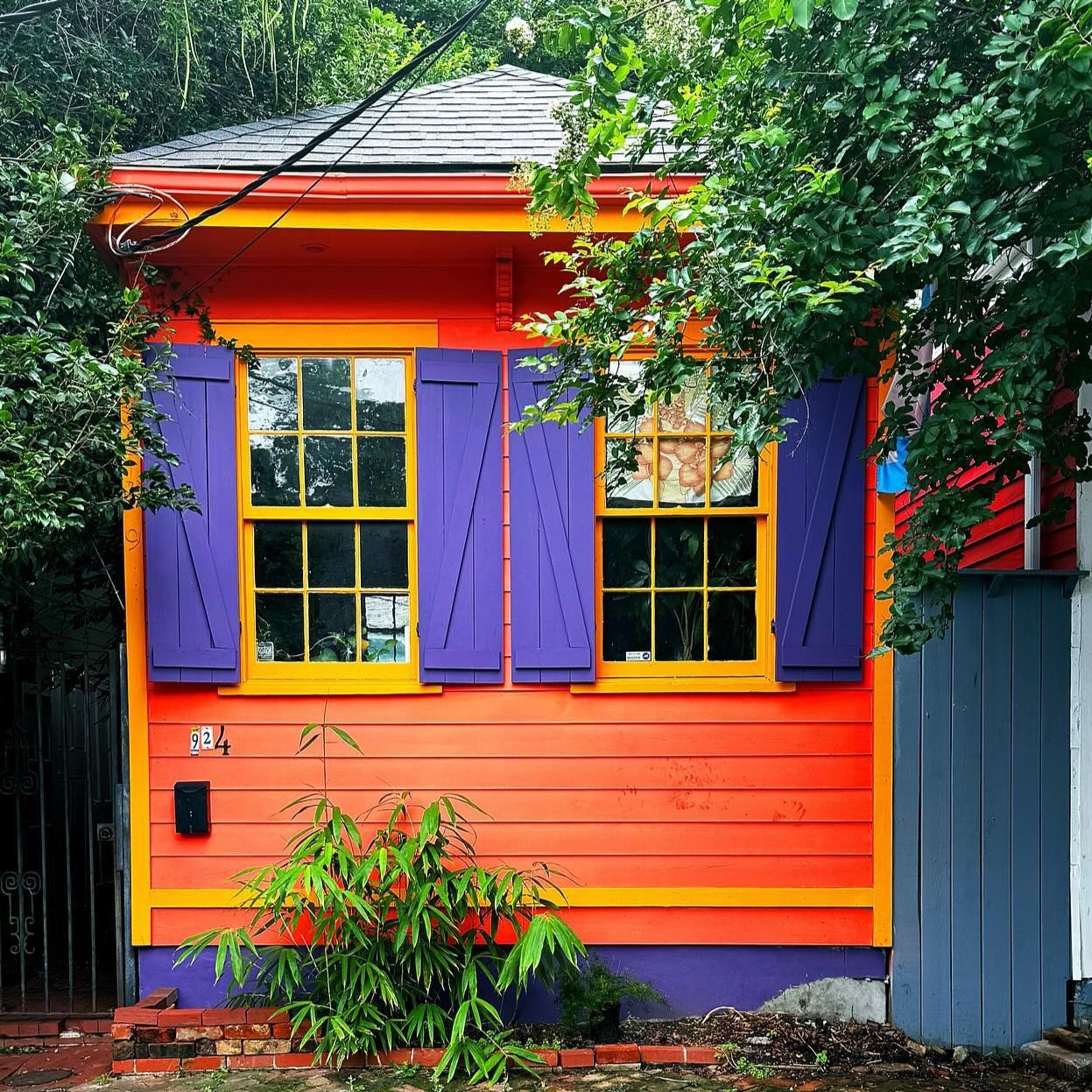 Small wooden house painted in bright orange, purple, and yellow with purple shutters and green plants in front