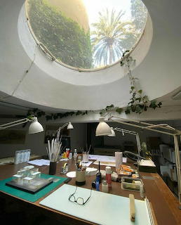 Artist studio with round skylight showing palm trees, desk with brushes, lamps, and art supplies