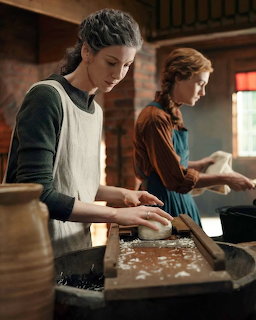 Two women in historical dress kneading dough in a rustic kitchen with pottery and wooden surfaces