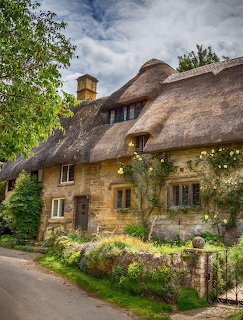 Stone cottage with thick thatched roof surrounded by colorful flower garden and greenery