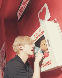 Young man with blonde hair leaning toward a red emergency box with a teddy bear inside labeled In Case of Falling in Love