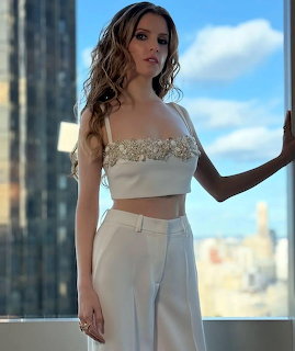 Anna Kendrick standing by a window in a white outfit with a blurred city view behind her