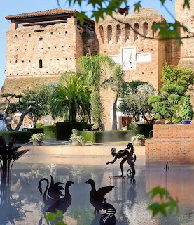 Medieval Italian castle with brick walls, arched entrance, fountain with bronze swan sculptures, surrounded by palm trees