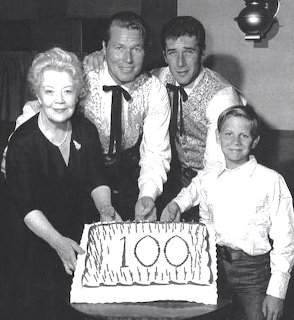Four people in vintage western attire posing behind a cake reading 100